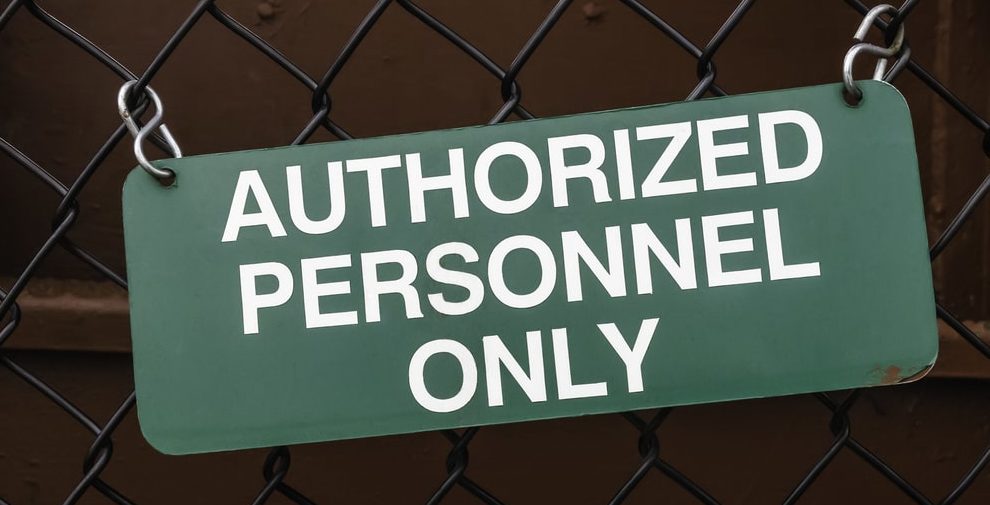 Light green outdoor sign, slightly crooked, on black chain-link fence by dark red steel wall AUTHORIZED PERSONNEL ONLY